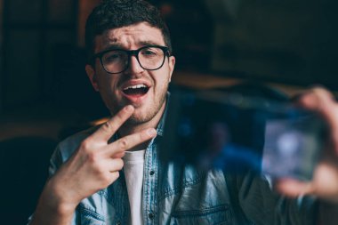 Heyecanlı erkek blogcu barış parmaklarını gösteriyor ve kapalı mekanlarda komik fotoğraflar çekiyor. Genç duygusal hippi, akıllı telefon cihazıyla wifi 'ye bağlı sosyal ağlar için fotoğraf çekiyor.