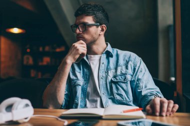 Kafası karışmış hipster çocuk kendi blogu için yazı yazmayı düşünüyor. Kapalı bir masada oturuyor, düşünceli erkek öğrenci sınava hazırlanıyor. Masaüstünde ders kitabıyla oturuyor.