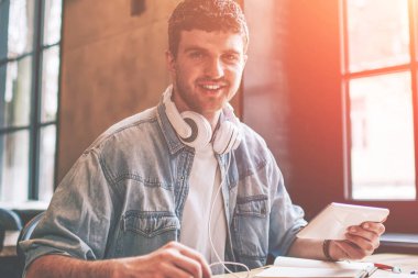 Elektronik kulaklıkla müzik dinlemek için medya uygulaması kurarken, elinde dokunmatik pedi olan neşeli hipster adamın portresi. Sohbet için teknolojiyi kullanan pozitif adam.
