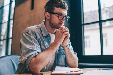Bulmaca çözme gözlüklü erkek yazar. Masaüstünde oturup makale için fikirler düşünüyor. Düşünceli, kafası karışık hipster çocuk, ders için hazırlanıyor.