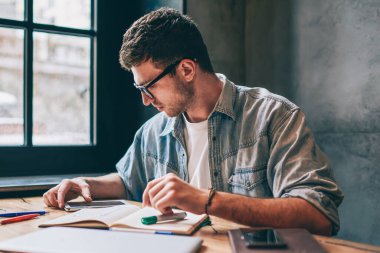 Wi-Fi bölgesindeki modern teknolojiyi kullanarak ders için araştırma yapan genç erkek öğrenci. Not defterine çizimler çizerken üniversite hakkında haber okuyan zeki hippi.