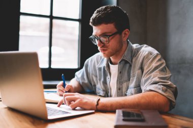 Teknoloji ve iletişim kavramı, gözlüklü zeki erkek kopyacı dizüstü bilgisayarda Wi-Fi 'ye bağlı, netbook ekranına bakan ve iş için notlar yazan genç adam.