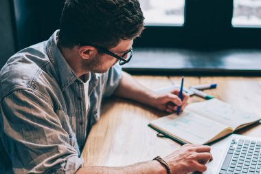 Gündelik giyinmiş hipster çocuk eğitim kitabında bir organizasyon planı yazıyor. Masaüstü bilgisayarında netbook 'la oturuyor, genç adam not defterine kompozisyon yazıyor ve modern teknolojiyle akademi sınavına hazırlanıyor.