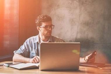 Yakışıklı erkek akıllı telefon cihazında profesyonel kontrol süresi ve içerideki bilgilendirme güncellemesi, sıradan hipster adam dizüstü bilgisayarın önünde oturuyor ve uygulamayla cep telefonu numarasını çeviriyor.