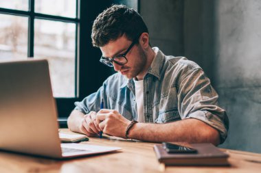 Düşünceli genç erkek grafik tasarımcısı dizüstü bilgisayarın önünde oturuyor, sıradan giyinmiş adam yeni fırsatlar arama olasılığını düşünüyor, serbest çalışan küçük işletme sahibi kapalı alanda şüphe duyuyor