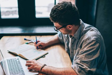 Gündelik giyinmiş hipster çocuk eğitim kitabında bir organizasyon planı yazıyor. Masaüstü bilgisayarında netbook 'la oturuyor, genç adam not defterine kompozisyon yazıyor ve modern teknolojiyle akademi sınavına hazırlanıyor.