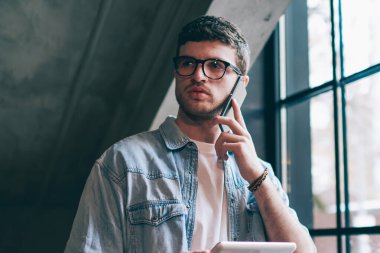 Gözlüklü düşünceli erkek kullanıcı göz koruması sağlıyor. Kapalı alandaki telefon görüşmelerini inceliyor. Konsantre hipster adam dokunmatik ped tutuyor ve cep telefonuyla müşteri hizmetlerini arıyor.