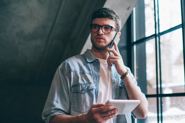 Gözlüklü düşünceli erkek kullanıcı göz koruması sağlıyor. Kapalı alandaki telefon görüşmelerini inceliyor. Konsantre hipster adam dokunmatik ped tutuyor ve cep telefonuyla müşteri hizmetlerini arıyor.
