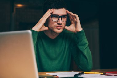 Ciddi bir erkek girişimci, elinde kağıtlar ve dizüstü bilgisayarla masa başında oturarak başlangıç projesi üzerinde kafa yoruyor, göz giysisi içindeki kafası karışmış genç bir öğrenci, derslerini düşünüyor ve kapalı alandaki çalışma sürecini planlıyor.