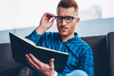 Skiled Kızıl saçlı genç adam portresi gözlük coworkin uzayda ilginç bir kitap okumak için sabit. Kahve dükkanında kapalı elinde ders kitabı ile oturan gözlük şık sahibi