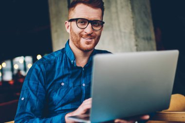 Portre pozitif sakallı erkek grafik tasarımcı Kızıl saçlı ve modern dizüstü bilgisayar, yeni proje üzerinde kablosuz 4 g için bağlıyken kameraya gülümseyen gözlük internet kafede