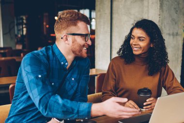Gülümseyen erkek öğretmen dizüstü bilgisayar coworking içinde oturan neşeli Afro-Amerikan öğrenci çalışma için yeni konu açıklama Kızıl saçlı. İki hipsters yeni proje işbirliği sırasında tartışıyor