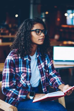 Zeki Afro-Amerikan öğrenci kıvırcık saçlı edebiyat kitap kütüphane kullanarak seminer için hazırlanıyor. Boş ekran alanı ile modern netbook. Koyu tenli teen uzakta arıyorum elinde ders kitabı ile
