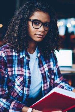 Kıvırcık saçlı ve kitap okuyan ile Kitaplığı'nda otururken kameraya bakarak gözlük güzel akıllı Afro-Amerikan öğrenci portresi. Zeki koyu tenli teen elinde ders kitabı ile