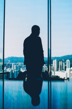 Büyük cam pencere duran ve şaşırtıcı akşam toplayan modern şehir gökdelenler ve yüksek mountaons ile arka plan üzerinde zor iş günü sonra seyir şirketinin sahibi silüeti geri