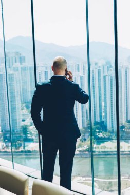 Ofiste ayakta ve metropolis harika manzarasını penceresinden bakarak ssuccessful iş projesi sonuçlarını smartphone aygıt yolu ile söz formal giyim erkek girişimci arkadan görünüm