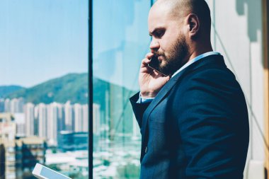 Resmi giyim hareket çek hesabı sırasında smartphone ile modern tablet duran cam pencere arka plan üzerinde yüksek gökdelenler ile yakınındaki bankacılık hakkında konuşmak emin gurur CEO'su yan görünüm giymiş