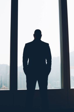 Arkadan görünüşü şık giyim zor bir gün sonra çalışma konuları üzerinde düşünürken pencereden dışarı bakarak gurur CEO'su giymiş erkek. Kapalı duran kendi şirket ofisinde işadamı silüeti