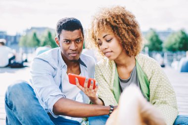 Gündelik giyinmiş Afrikalı Amerikalı çift dışarıda dinleniyor ve blogda video izlemek için modern akıllı telefonu kullanıyor. Koyu tenli gençler telefonda uygulamayı güncelliyor.