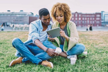 Mutlu çift birlikte parkta yeşil çimlerde vakit geçirir ve modern dokunmatik ped cihazında video izlerler. Afro-Amerikalı genç adam ve kadın tablet üzerinde birlikte alışveriş yapıyorlar.