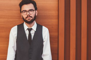 Şık giyimli bir adamın yarım boy portresi. Reklam alanında duvarın yanında duruyor ve boş zamanlarında kameraya bakıyor. Kendine güvenen, moda yelekli genç adam boş zamanın tadını çıkarıyor.