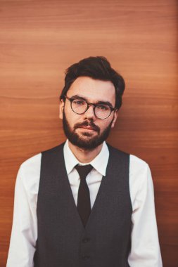 Gözlüklü, beyaz sakallı hippi, sokak duvarının yanında duran ve kameraya bakan, reklam alanına yakın resmi yelek giyen, kendine güvenen genç bir adamın yarım portresi.