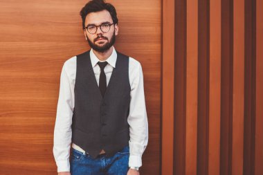 Şık giyimli bir adamın yarım boy portresi. Reklam alanında duvarın yanında duruyor ve boş zamanlarında kameraya bakıyor. Kendine güvenen, moda yelekli genç adam boş zamanın tadını çıkarıyor.