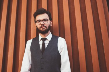 Beyaz tenli, resmi giyinmiş bir erkek öğrenci şehir caddesinde duvarın yanında dikilirken kameraya bakıyor. Yelekli genç hippi, açık hava alanında boş zaman harcıyor.