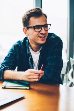 Genç gülümseyen zeki freelancer kahve dükkanında vakit zevk serin gözlük yakışıklı. Ahşap masa kafede oturan olumlu hipster adam. Mutlu adam çalışmaları sırasında kapalı eğleniyor