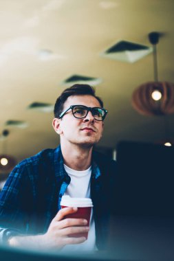 İyi görünümlü yetenekli öğrenci trendy Eyewear hazırlık kurs için mola sırasında lezzetli kahve zevk. Take away içecek kapalı masada otururken yakışıklı hipster adam