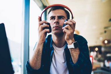 Kapalı tahta masada oturan yeniden oluştururken büyük modern kulaklık alarak genç yakışıklı durulması erkek öğrenci. İyi görünümlü bir adam baş aygıt kafede favori müzik dinleme için koyarak