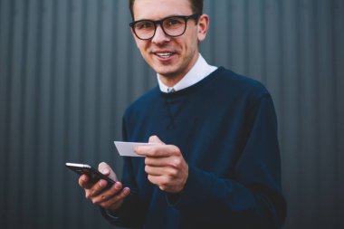 Yarım uzunlukta trendy Eyewear açık havada eğlence keyfini gülümseyen yakışıklı erkek öğrenci portresi. Kamera tutarken bakarak neşeli hipster adam ziyaret kart ve akıllı telefon yolu ile online rezervasyon