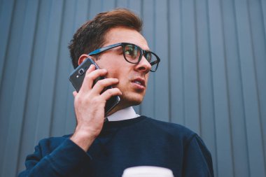 Yakışıklı dalgın hipster adam trendy Eyewear kentsel ayarında alan reklam ile duvar yanındaki yürüme. Promosyon arka plan karşı duran cep telefonunda konuşan genç iyi görünümlü adam