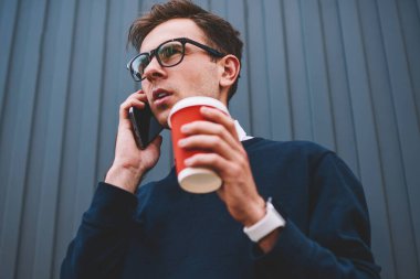 Yakışıklı hipster adam smartphone uygulama ve Internet kullanarak mobil konuşmayı gündelik kıyafeti giymiş. Ayarlama ve telefonda konuşurken take away kahve ile Caddesi'nde yürüyen genç adam