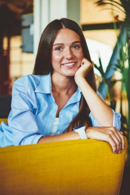 İçerde oturan, boş zamanlarında eğlenen, rahat giyinen, gülümseyen ve kampüste arkadaşını beklerken kameraya bakan beyaz, başarılı bir kadının portresi.