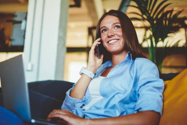 Pozitif başarılı hipster kızı cep telefonundaki bakiyeyi kontrol etmek için arıyor. Bilgisayarla kapalı alanda dinlenirken modern teknoloji ve iletişim kavramını kullanıyor.