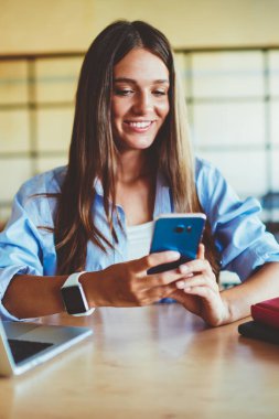 Modern cep telefonu ve içerideki 4G bağlantısını kullanarak arkadaşlarıyla sohbet eden mutlu bir kadın, akıllı telefon cihazına SMS mesajı yazarken bilgisayarının başında oturan pozitif hipster kızı.