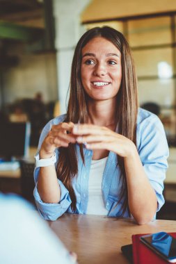 Olumlu gülümseyen kadın, esmer saçlı, ahşap masada oturan ve erkek arkadaşıyla önde konuşan, başarılı, beyaz giyimli, sıradan giyinmiş, erkeklerle vakit geçiren ve fikirleri tartışan bir kız.