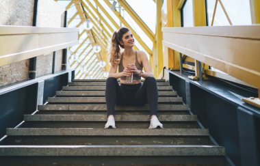 Spor elbiseli genç bir kadın açık havada merdivenlerde oturuyor, elinde su şişesi, ve gülümsüyor. Sarı kirişlerin parlak geometrik arkaplanının çerçevelediği bir egzersizden sonra rahatlamış görünüyor.