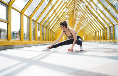 Genç bir kadın, sarı kirişlerle modern cam bir koridorda uzanıyor, akıllı bir telefon tutuyor ve müzik dinliyor, hareketli, güneşli bir kentsel alanda egzersiz yapmaya hazırlanıyor.