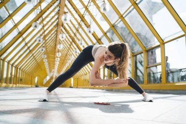 Genç bir kadın, kolları dizinin üzerinde katlanmış, parlak, geometrik bir koridorda geniş bacaklı bir esneme hareketi yapıyor. Kulaklıkları takıp, yere yakın bir yerde telefonu olan esnekliğine odaklanıyor.