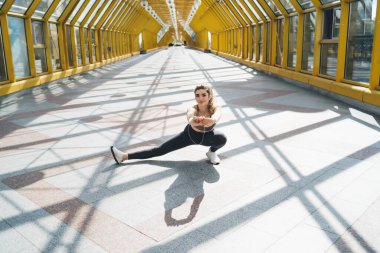 Genç beyaz kadın şehir ortamında derin hamleler yapıyor. Spor sutyeni ve tayt giymiş, sabah antrenmanı sırasında esnekliğe odaklanan gülümsemeler, hareketli ve aktif yaşam tarzının tadını çıkarıyor