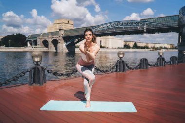 Güneşli bir günde nehir kenarında kartal pozuyla yoga yapan genç bir kadın. Odaklanmış, kolları birbirine dolanmış bir şekilde tek bacağı üzerinde dengede duruyor. Manzaralı bir köprü ve şehir zeminine karşı.