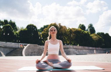 Genç bir kadın dışarıda nilüfer çiçeğiyle yoga yapıyor. Hafif bir gülümseme ile sakince oturur. Etrafı bulutlu bir gökyüzünün altında ağaçlarla çevrilidir. Refah temaları için ideal