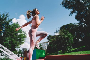 Hareket halindeki genç bir kadın dışarıda diz egzersizi yapıyor. Enerji ve zindeliği somutlaştırıyor. Parkta parlak, güneşli bir gün. Arka planda yeşil ve mavi gökyüzü var. Şık bir giysi giymişti.