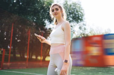 Akıllı telefon ve su şişesi tutan, spor sahasında renkli arka planda duran zinde genç bir kadın. Spor kıyafetleri giymiş, kendine güvenen ve açık hava ortamında motive olmuş.