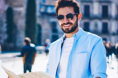 Siyah güneş gözlüklü mutlu sakallı genç adamın yarım boy portresi. Eski mimari şehrin kentsel ortamında dolaşırken kameraya gülümsüyor. Şovlarda ve doğru yönde harita kullanıyor.