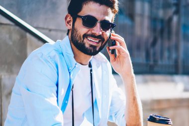 Mutlu sakallı hippi erkek arkadaşıyla ele gider için smartphone sittting kahve ile sokakta üzerinde gezici komik telefon konuşma yaparken kameraya gülümseyen şık güneş gözlüğü portresi