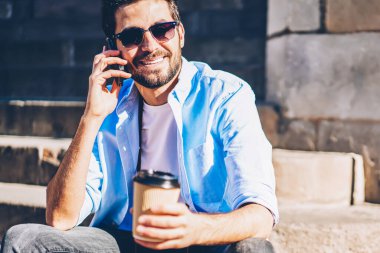 Güneş gözlüklü, gülen, akıllı telefonlu arkadaşlarıyla cep telefonuyla konuşan, elinde kahveyle merdivenlerde oturan neşeli erkek fotoğrafçı.