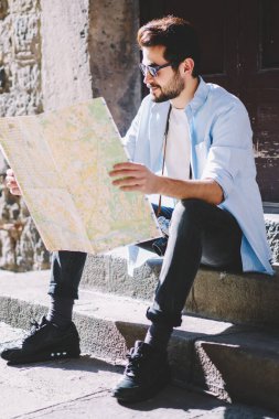 Düşünceli sakallı genç turist günlük kıyafetlerini giymiş seyahat haritasını dikkatle okuyor ve şehir merkezindeki merdivenlerde otururken bir sonraki gösteri için uygun rotayı belirliyor.